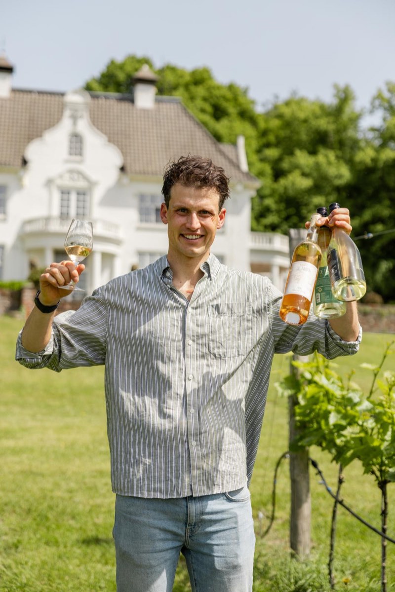 Rob met een glas en drie flessen wijn van De Tangh in de wijngaard, met het landhuis op de achtergrond